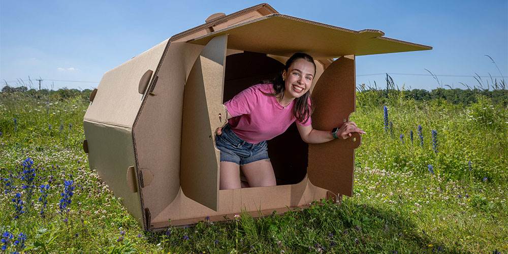 The image shows a tent made out of cardboard. It is being used for events. There is a woman inside the cardboard tent, opening the doors and looking outside.