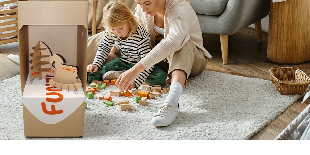 A mother is sitting with her son on the carpet and playing with toys. The funzy cardboard box is on the left side.